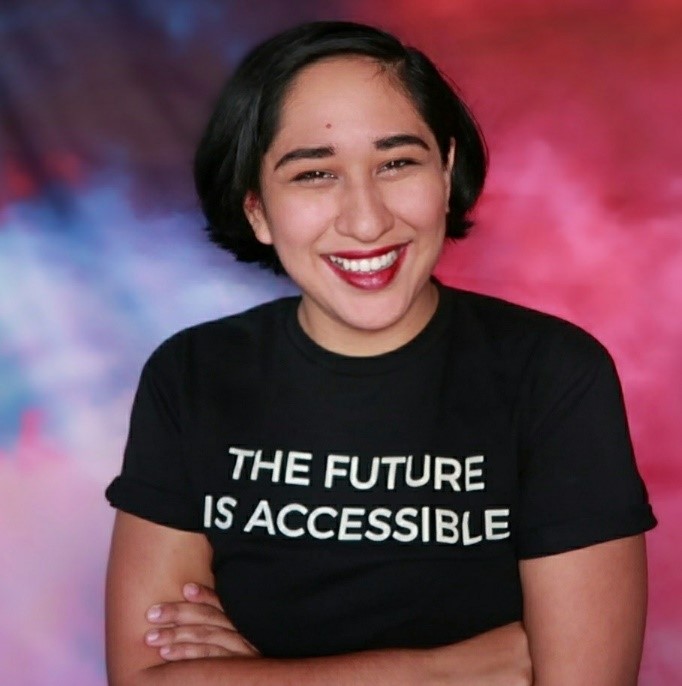 Image of Annie Segarra. A Latinx individual with short black hair. Arms are crossed in this photo and the shirt is black and says "the future is accessible."
