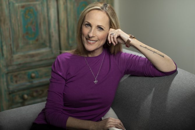 Image of actress and activist Marlee Matlin. A woman with blond straight hair sitting on a sofa in a purple shirt.