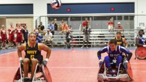 2 male individuals in wheelchairs racing towards an airborne volleyball in a competition.
