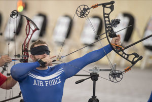 A male archer wearing a blue long sleeved shirt and a blind fold preparing to take a shot with his bow and arrow.