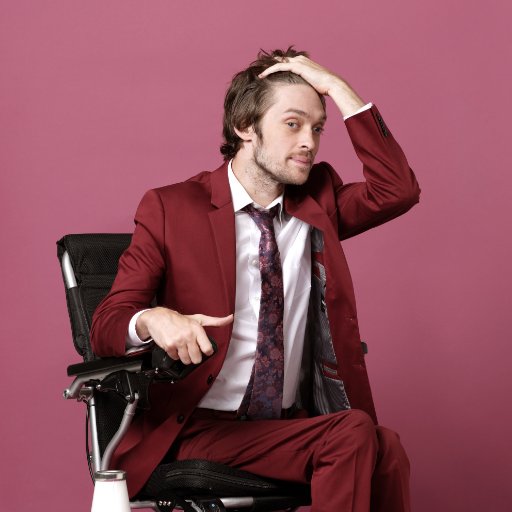Image of Zach Anner an author and comedian. He's in a red/maroon suit sitting in his wheelchair and running his hand through his hair.