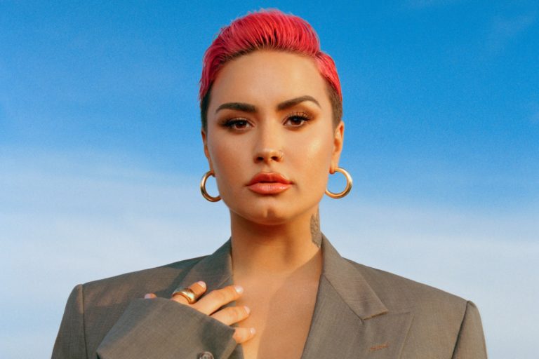 Image of Demi Lovato with close cropped pink hair wearing a professional jacket and a blue background.